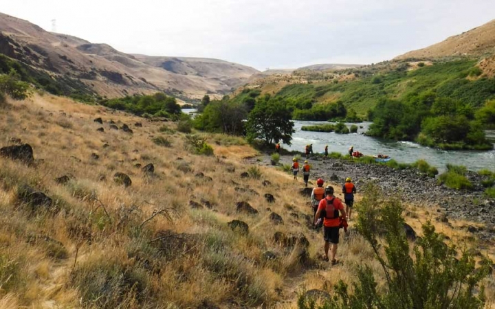 a group of students walk through a grassy and rocky shore, toward a blue river